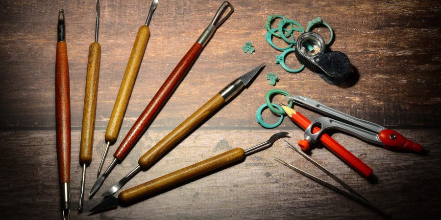 Prototyping tools, steel options, pointed pieces, multi-colored jewelry and gemstones, green wax rings, tweezers, gemstone trays, compasses and a magnifying glass placed on a wooden table.